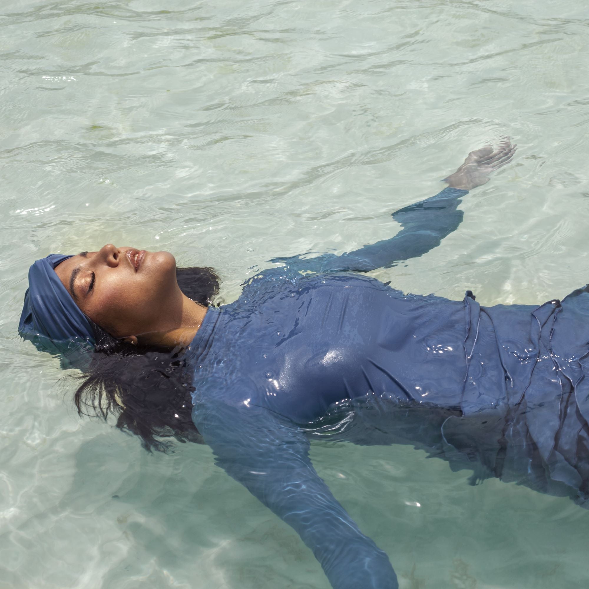 A woman floating in the sea wearing Lanuuk Lula swimwear in Azure and a matching Azure swim turban.